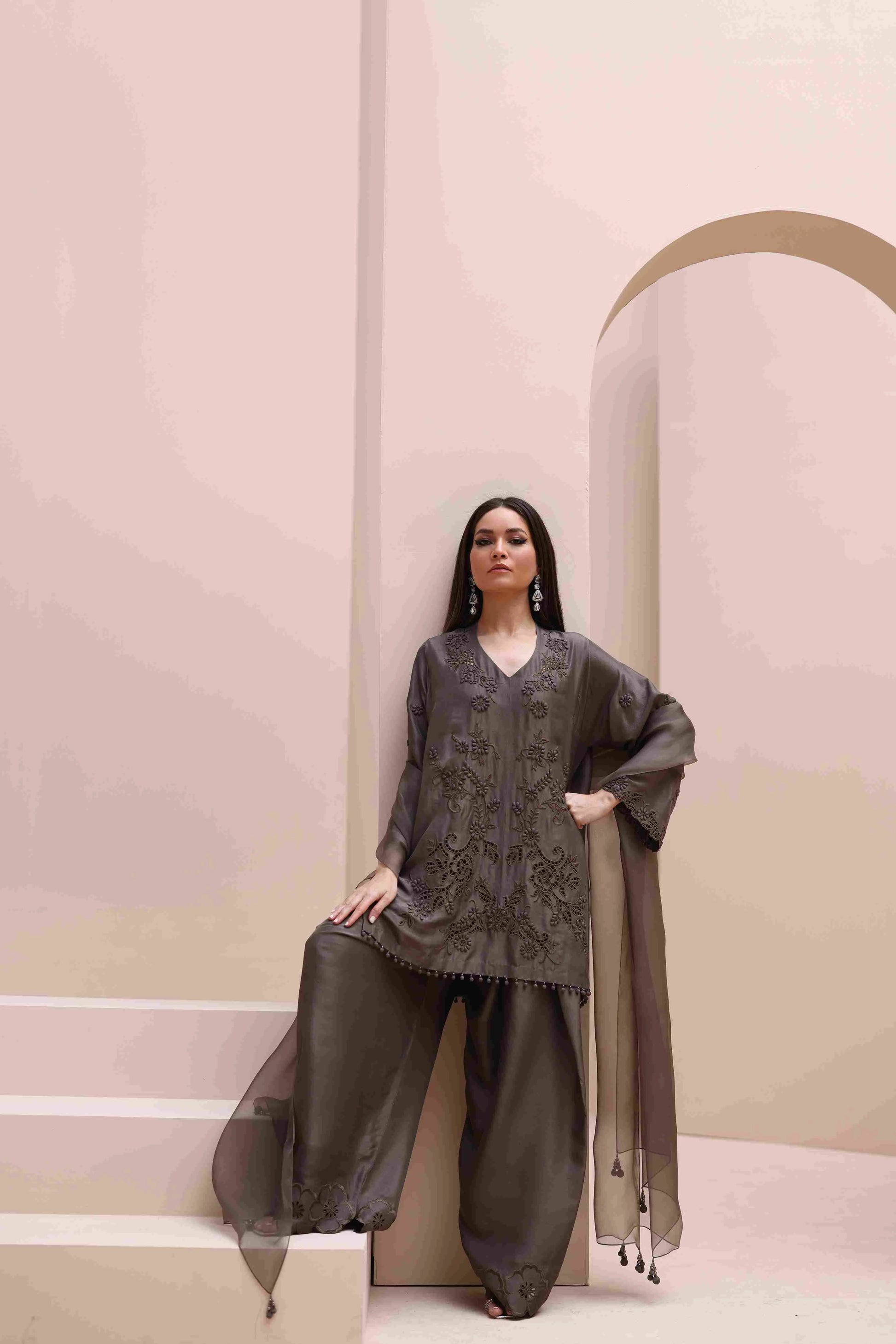 Woman in a dark gray traditional outfit standing against a pink and beige wall.