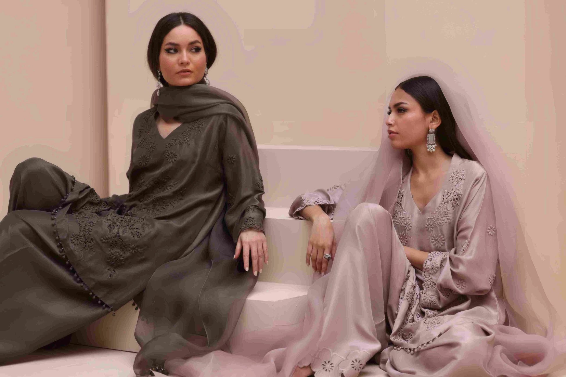Two women in traditional attire sitting on a couch against a light pink background