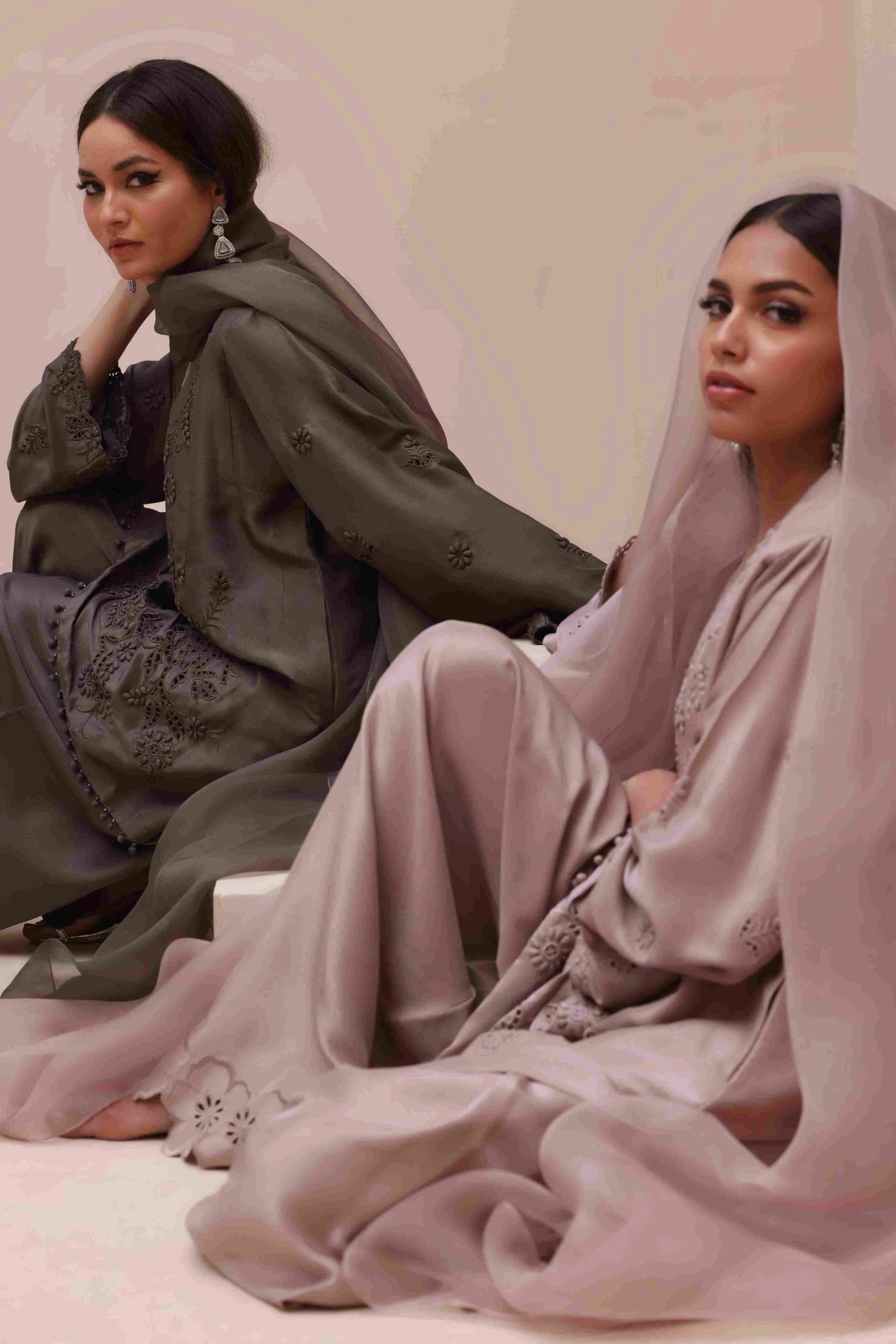 Two women in elegant dresses sitting on a pink surface with a neutral background