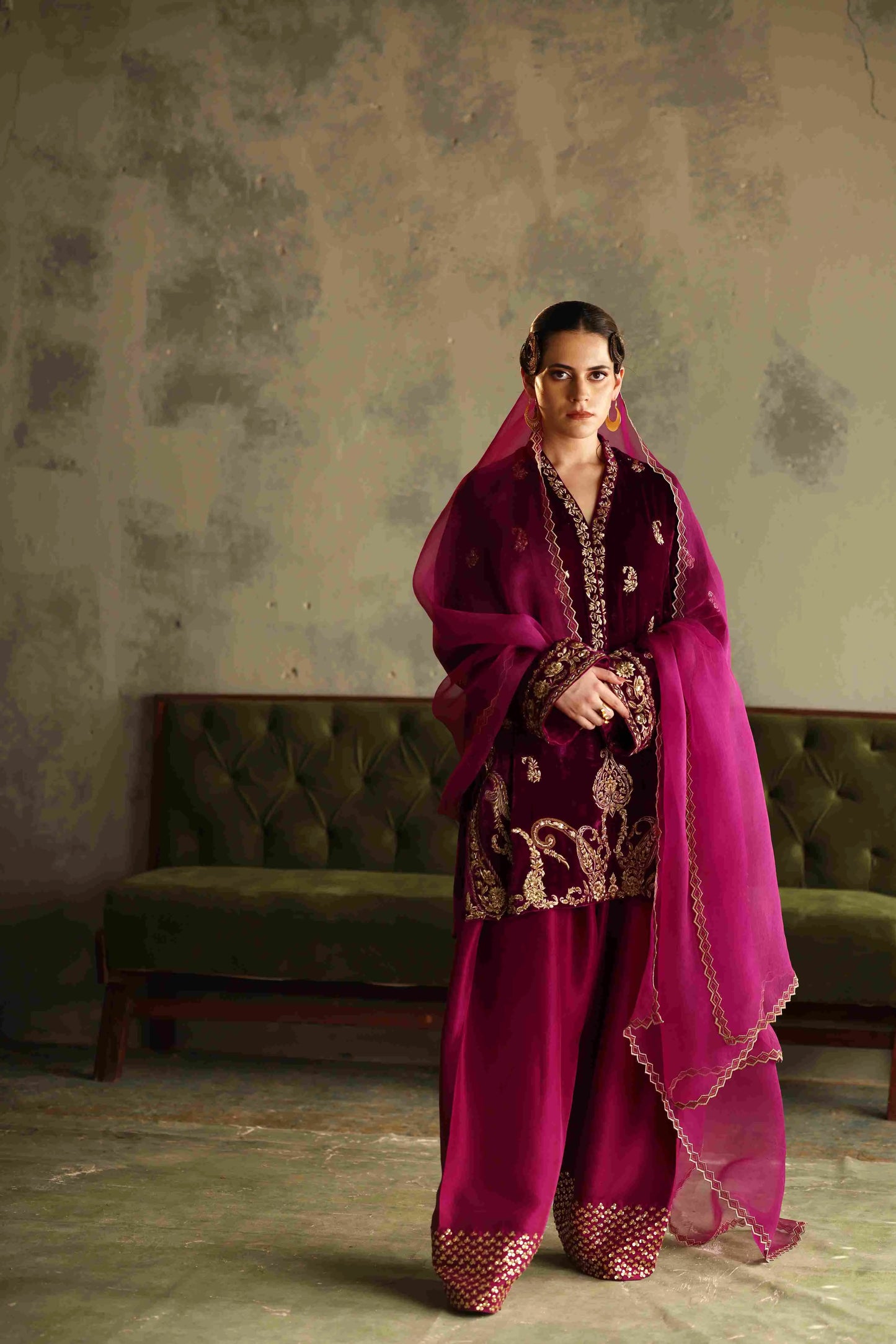 Woman in a traditional outfit with a pink dupatta standing in a room with a green sofa.
