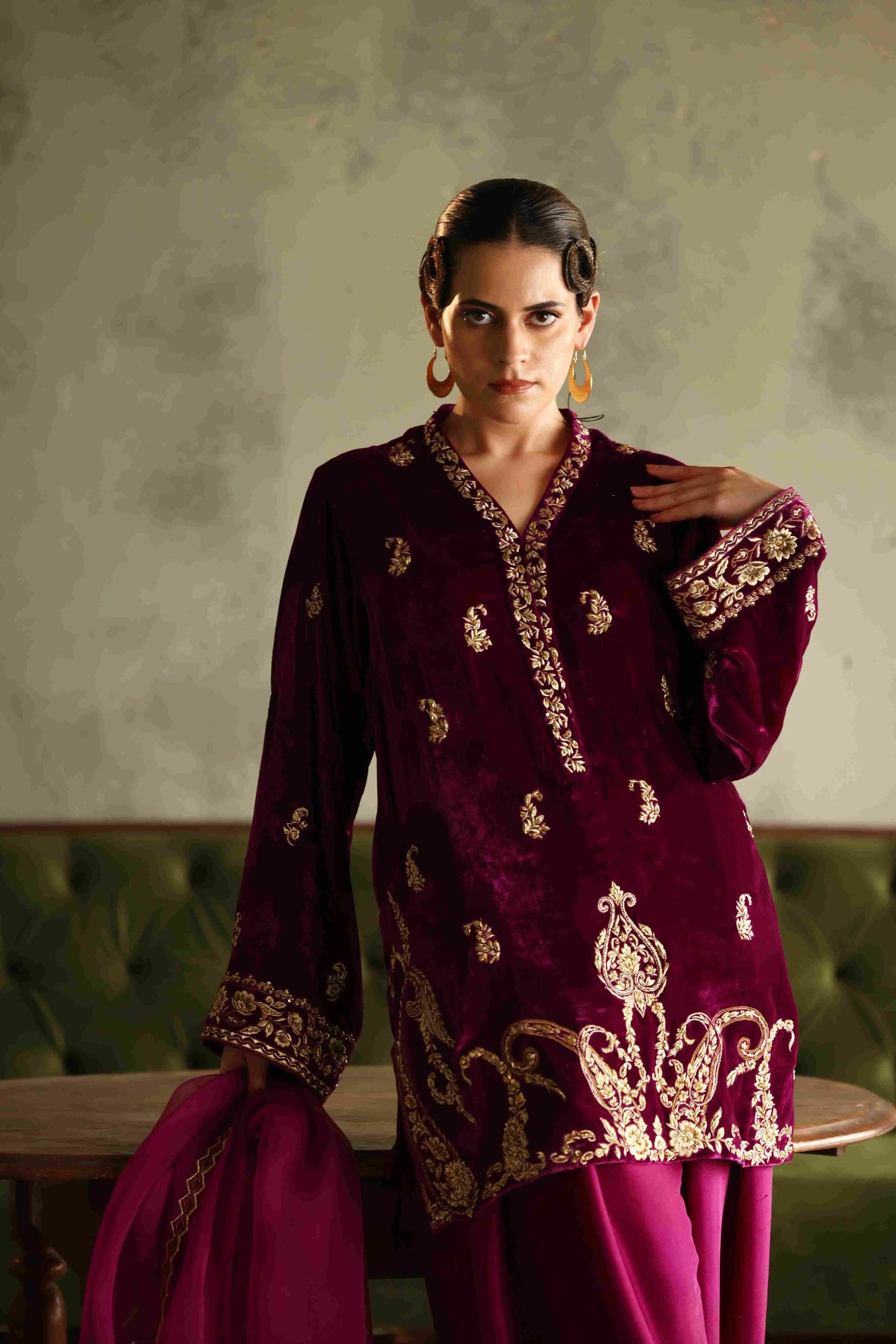 Woman wearing a maroon embroidered outfit with gold details against a green wall.