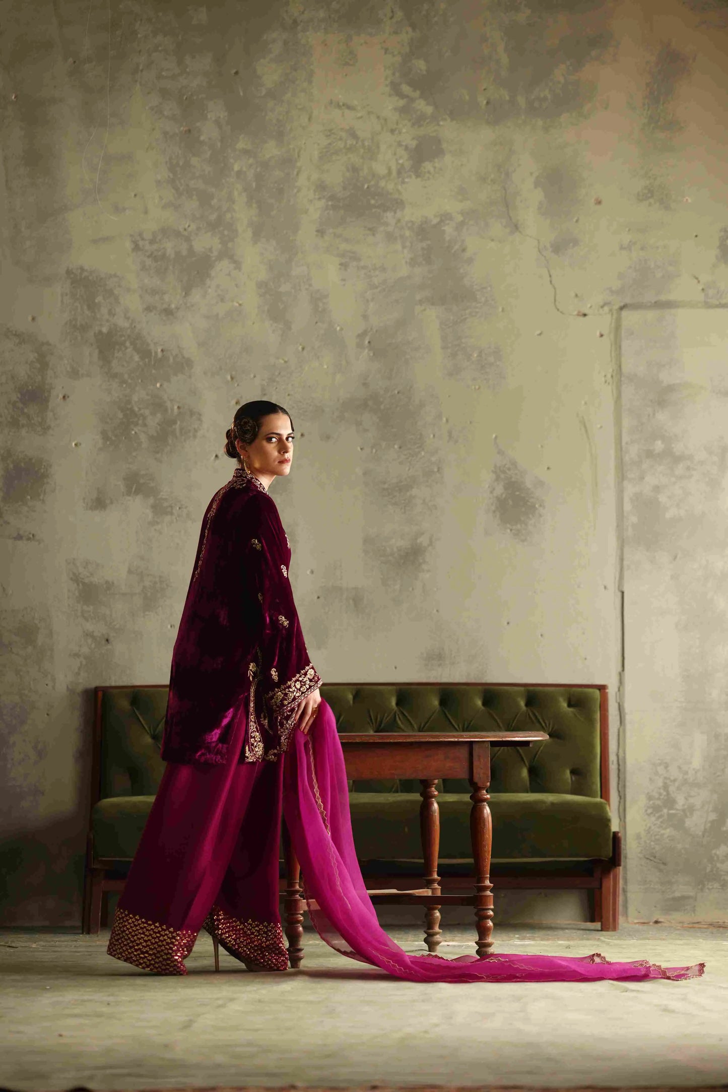 Woman in a vibrant pink saree with gold border standing against a textured wall.