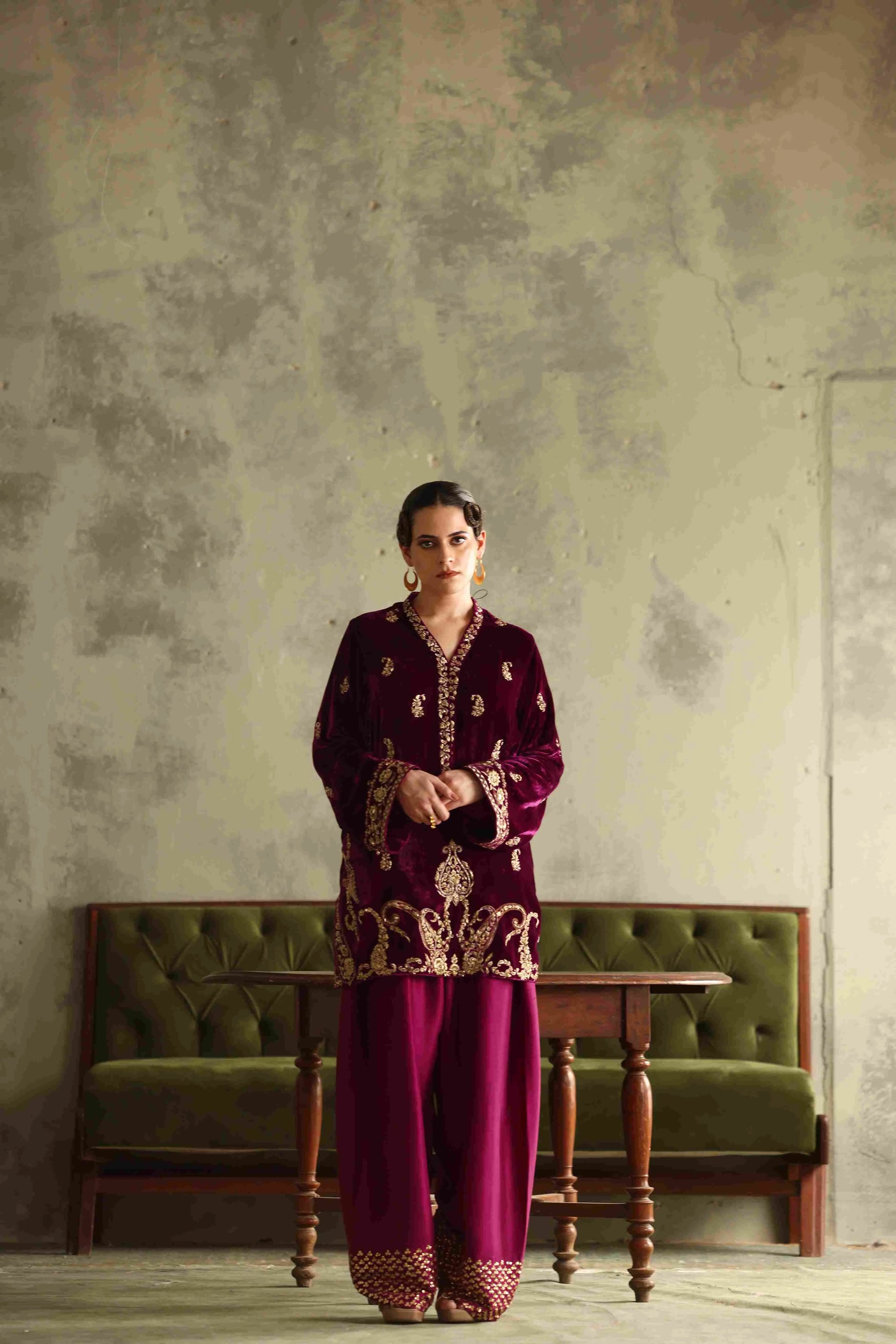 Woman in a burgundy embroidered outfit standing in front of a textured wall with a green sofa.