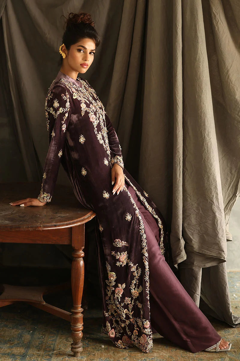 Woman in a dark purple embroidered outfit sitting on a wooden stool against a draped curtain background.