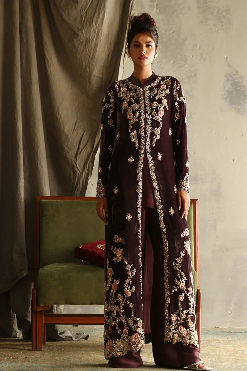 Woman wearing a dark purple embroidered outfit standing in a room with a green chair and gray curtain.