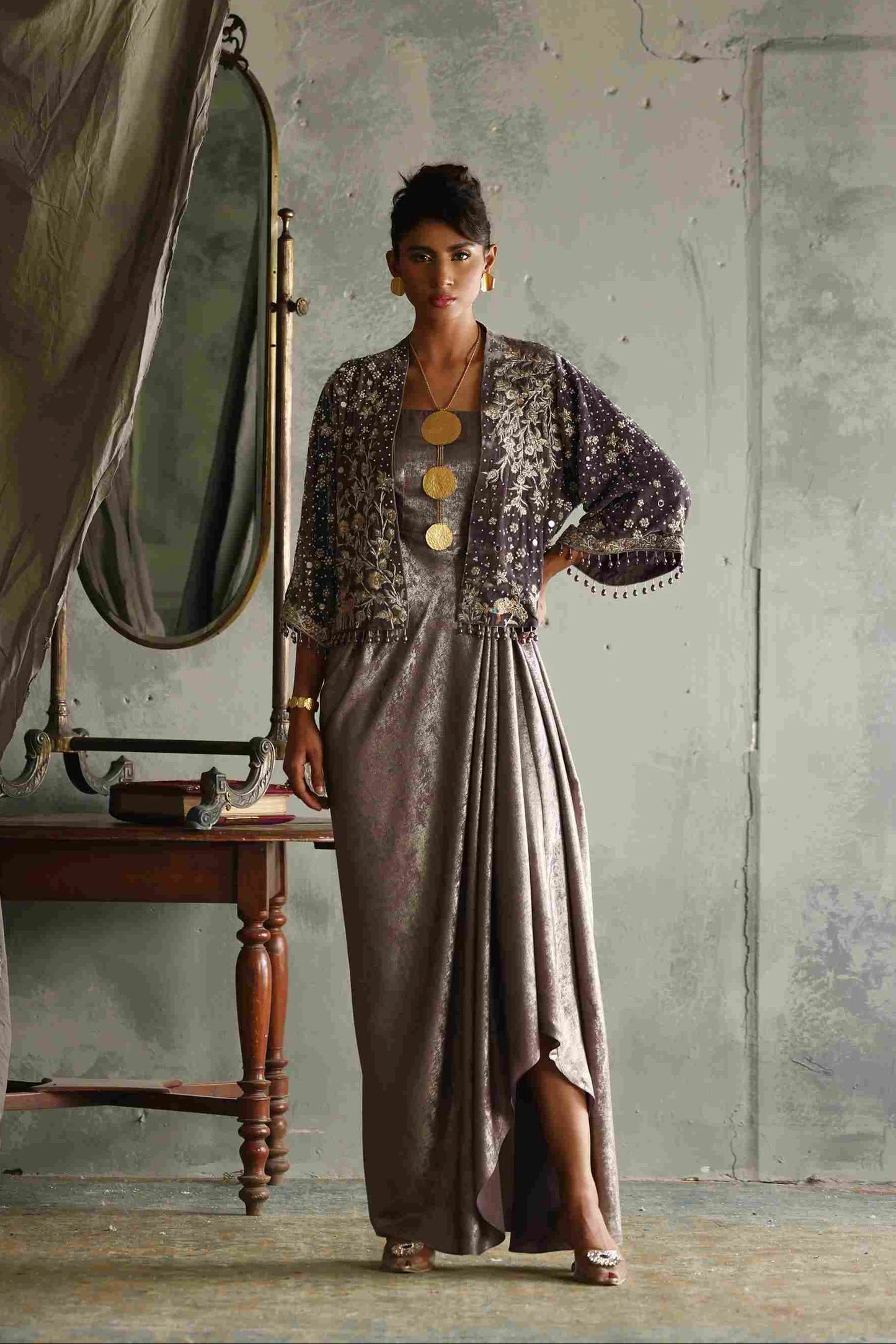 Woman in a long, elegant dress with a matching jacket standing in a room with a mirror and wooden table.