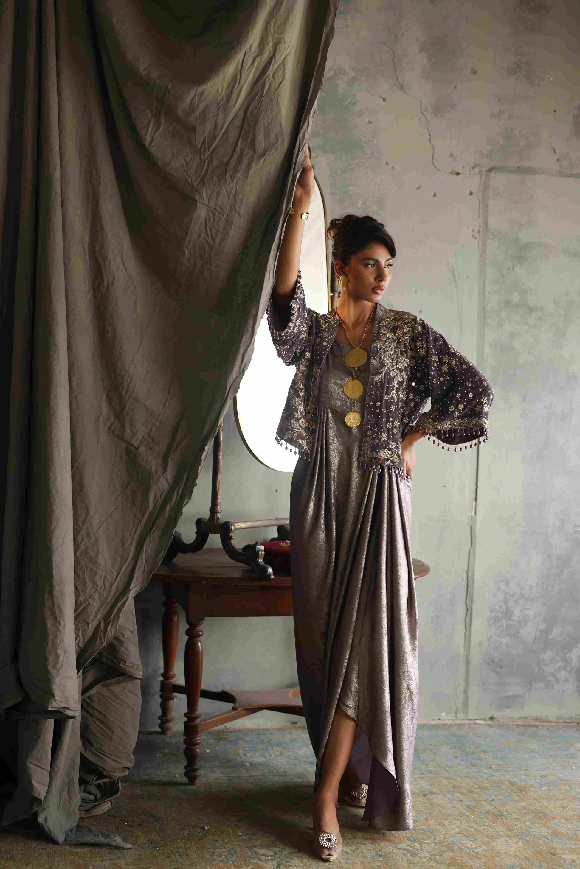 Woman in a long, flowing dress with a textured top standing in a room with a large curtain.