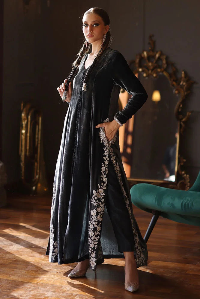 Woman in a long black dress with floral embroidery standing in a room with wooden floor and decorative mirror.