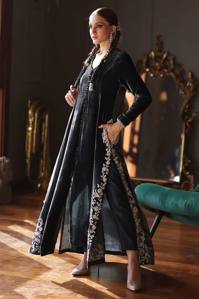 Woman in a long, dark coat with floral patterns standing in a room with wooden floors and a mirror.