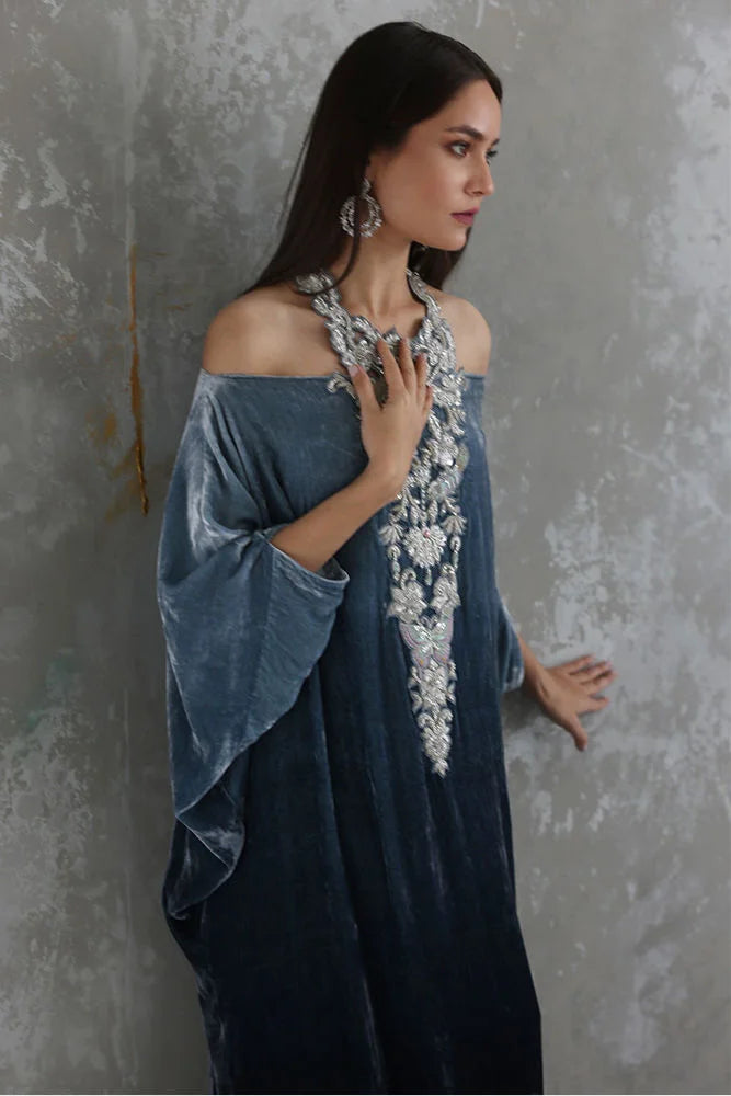 Woman wearing a blue off-shoulder dress with floral embroidery against a gray textured wall.