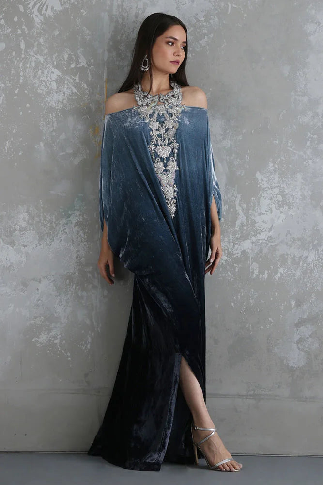 Woman wearing a blue evening gown with floral embellishments against a textured gray wall.