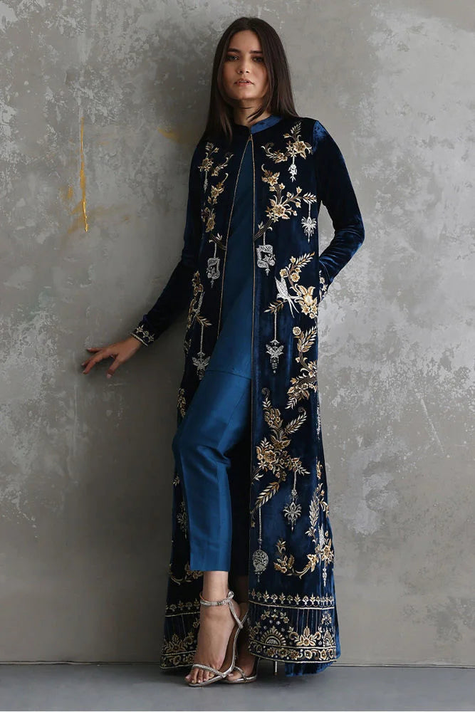 Woman wearing a long embroidered navy blue coat over a blue dress against a gray concrete wall.