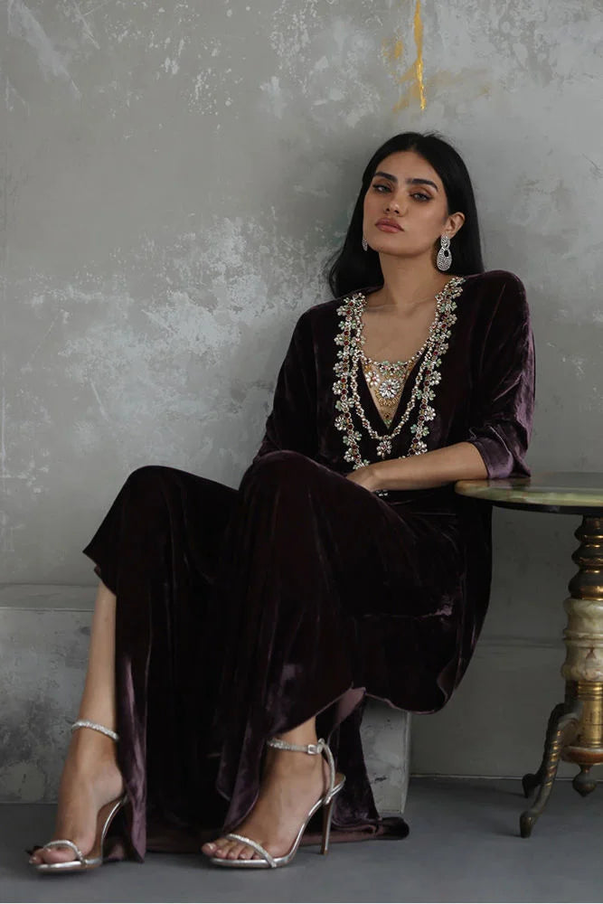 Woman in a dark purple outfit with intricate jewelry sitting against a gray wall.