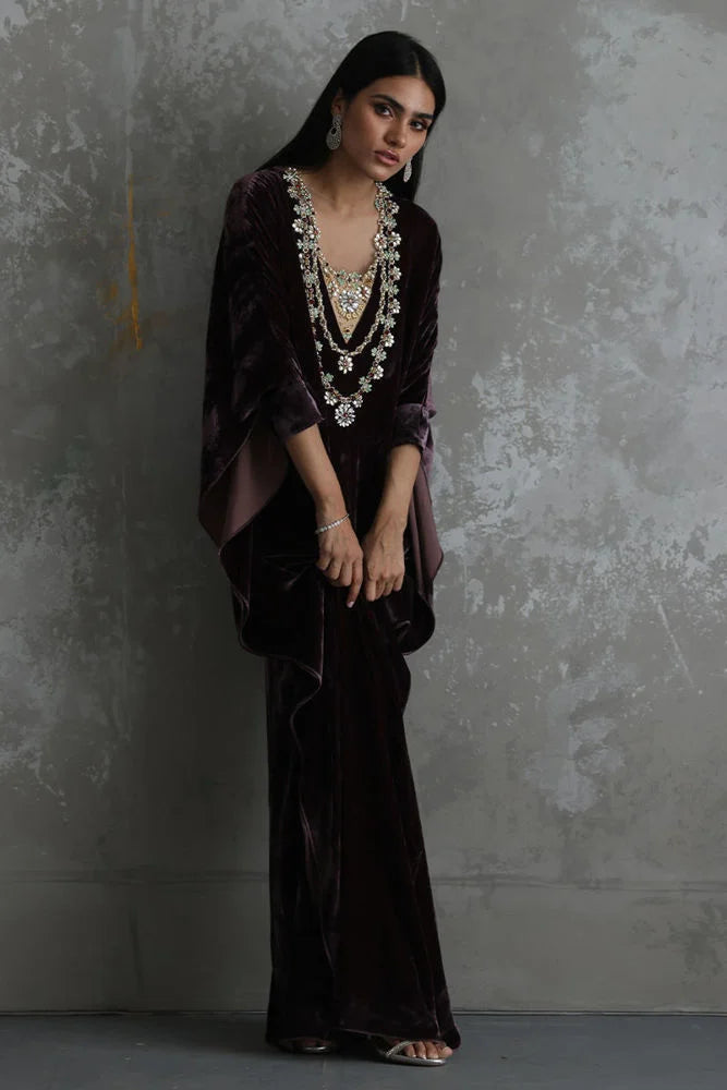Woman wearing an elegant dark purple gown with intricate detailing against a textured gray wall.