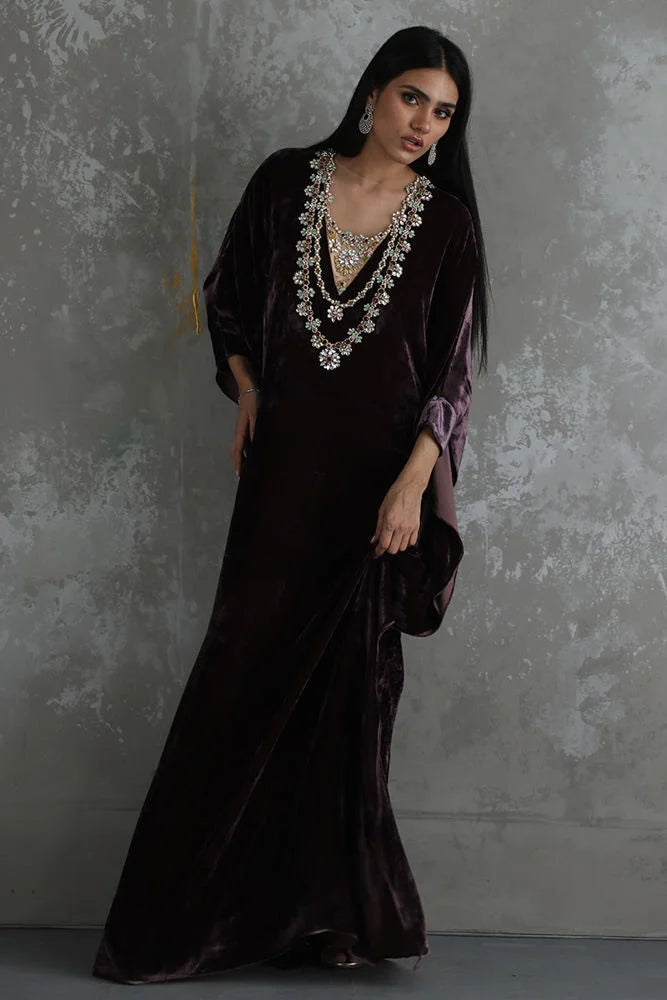 Woman in a long black dress with intricate jewelry against a textured gray wall.