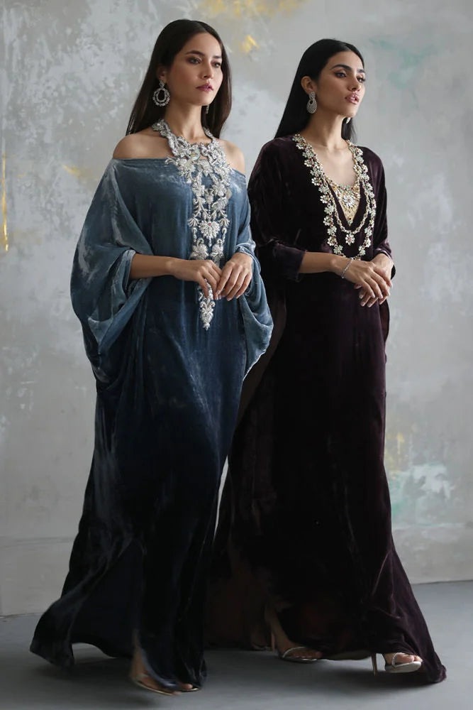 Two women in elegant evening gowns with intricate necklaces against a textured wall.
