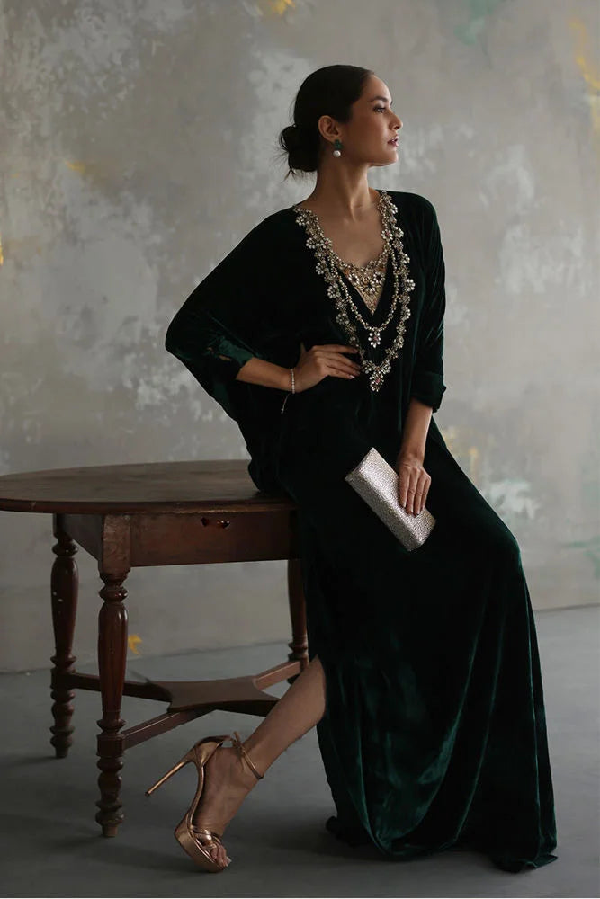 Woman in a dark green dress with jewelry sitting in a room with a wooden table and textured wall.