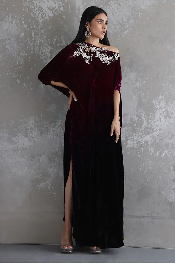 Woman wearing a dark red off-shoulder gown with floral embellishments against a gray textured wall.