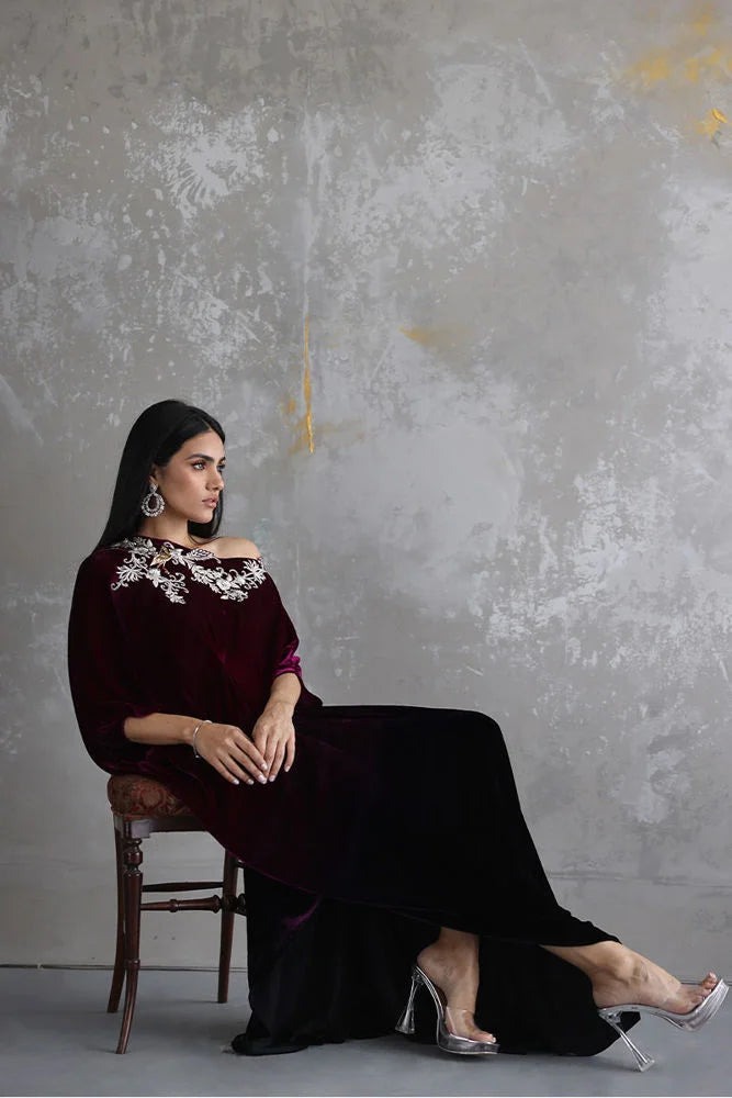 Woman in a dark purple dress with floral embellishments sitting on a chair against a textured gray wall.