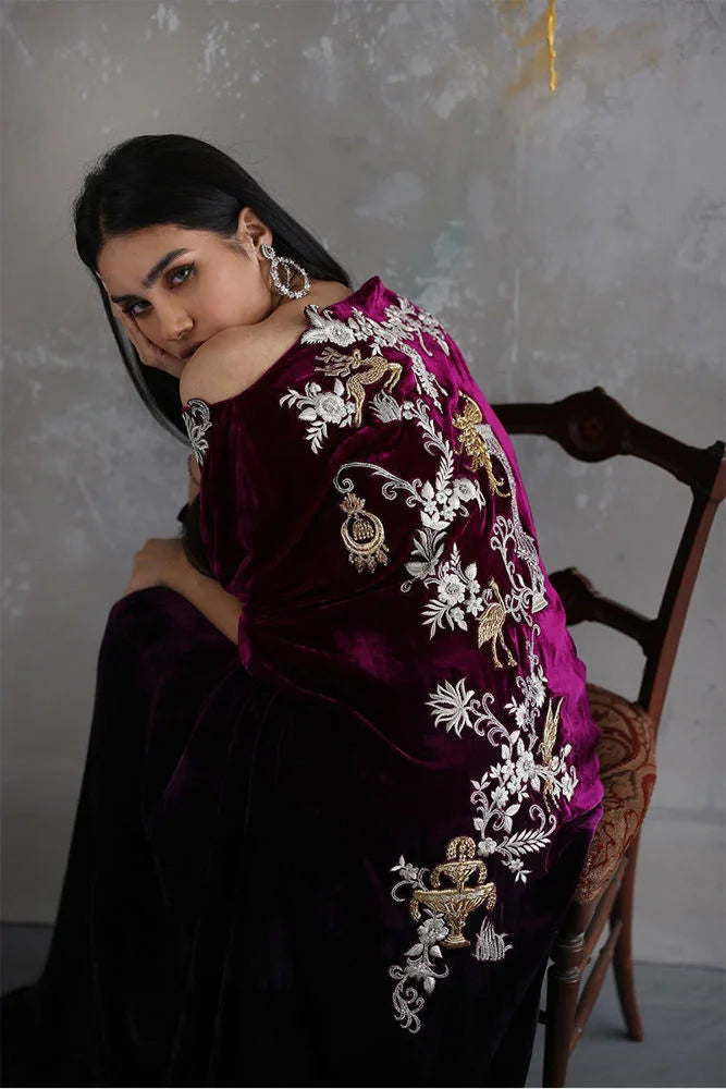 Woman wearing a dark purple traditional outfit with intricate white embroidery, sitting on a wooden chair.