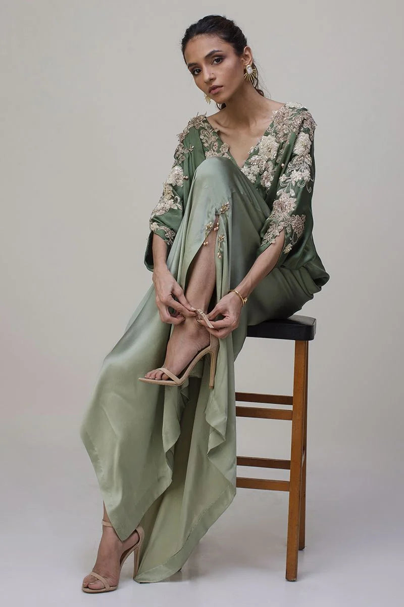Woman in a green dress with floral embroidery sitting on a wooden stool against a plain background