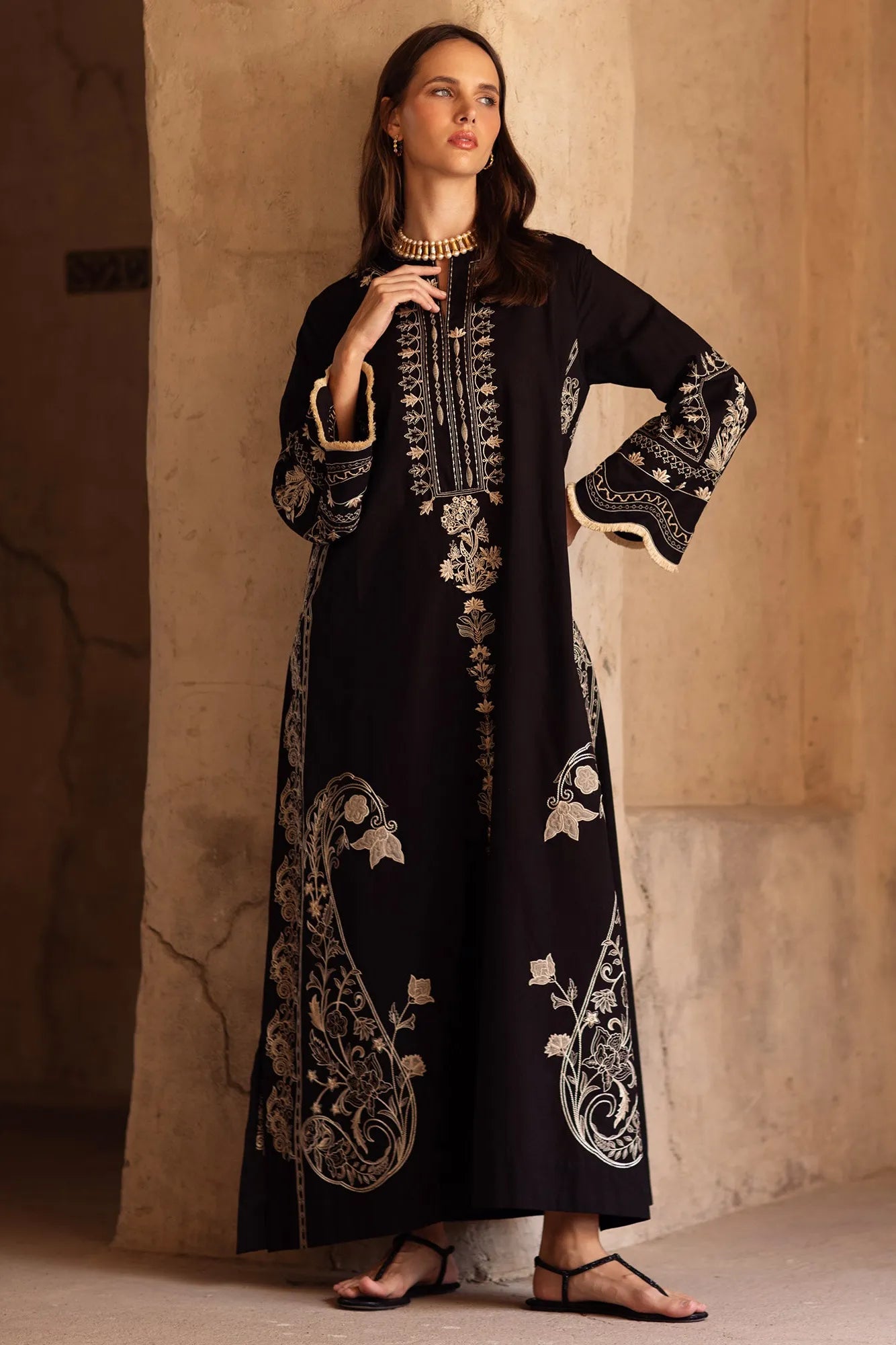 Woman wearing an embroidered black dress against a beige wall.