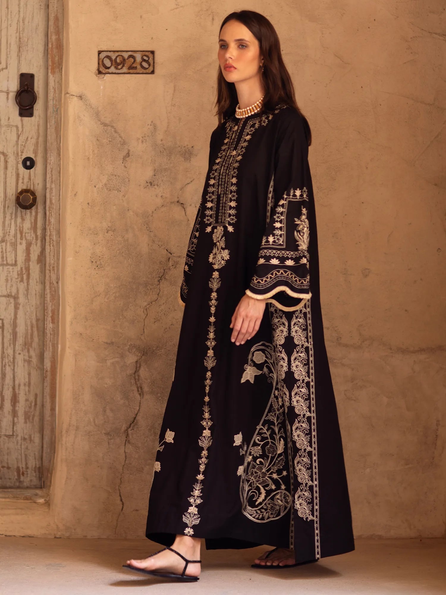 Woman wearing an embroidered black dress against a textured beige wall.