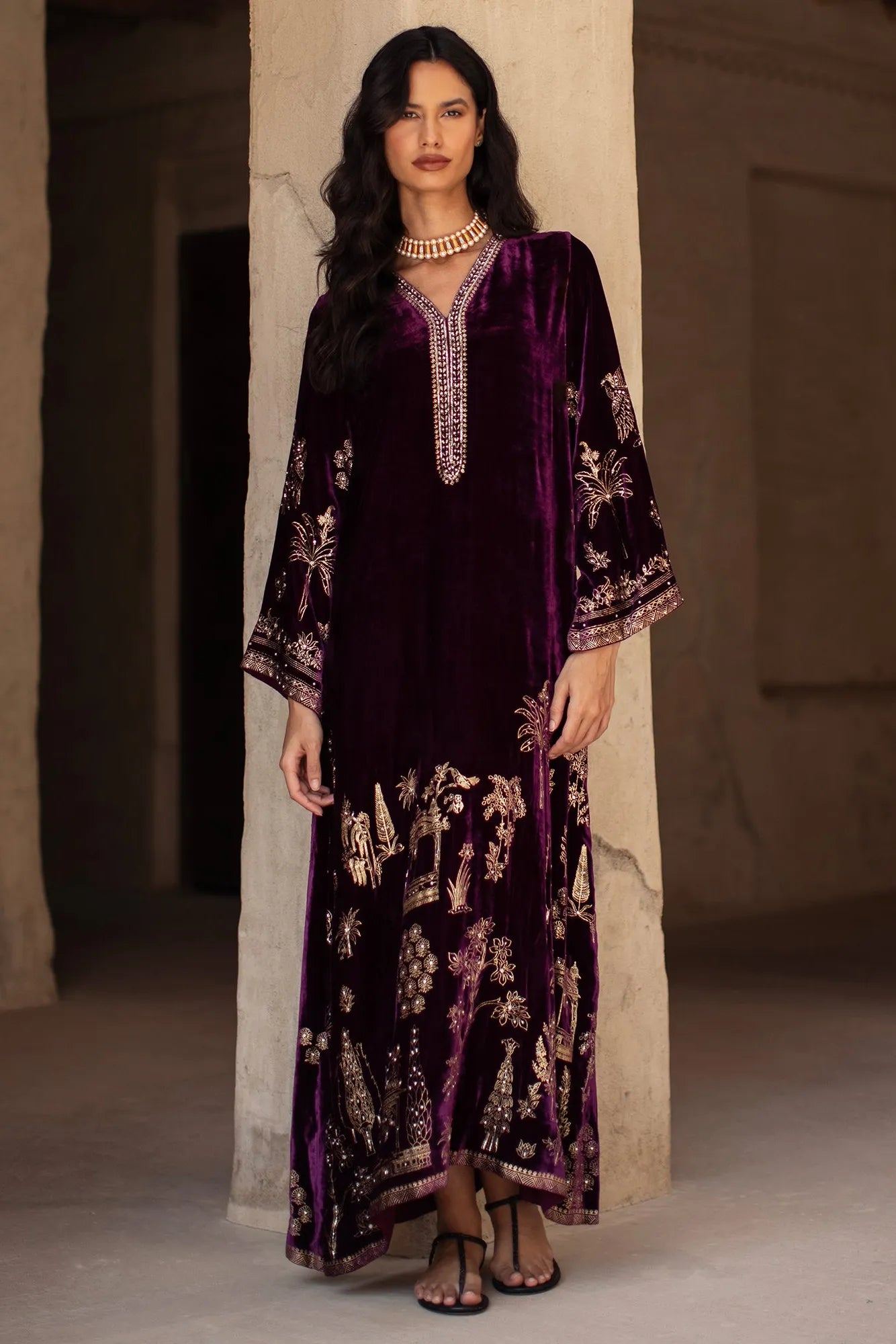 Woman wearing a dark purple embroidered outfit standing against a neutral wall.