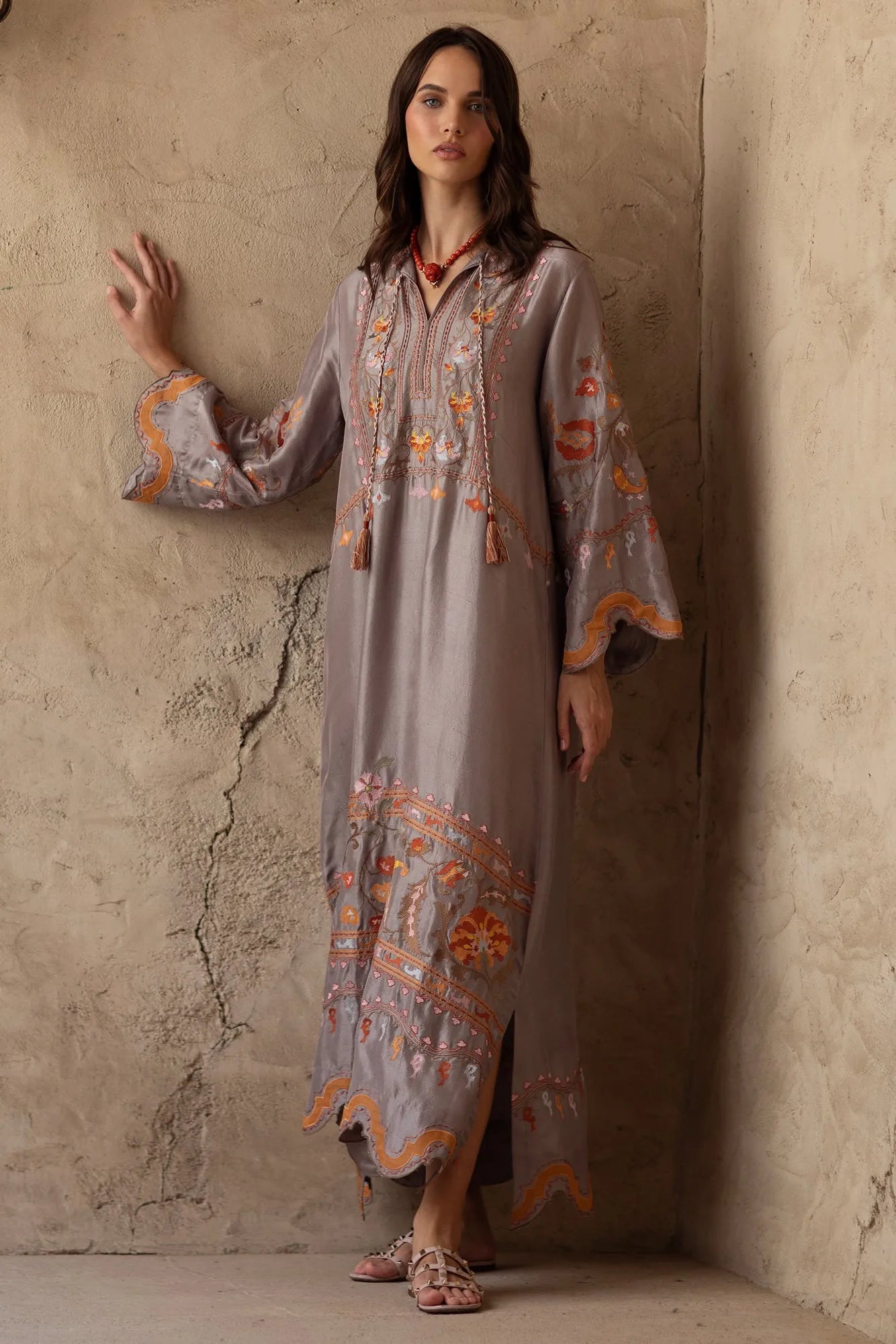 Woman wearing a long, embroidered dress against a textured wall.