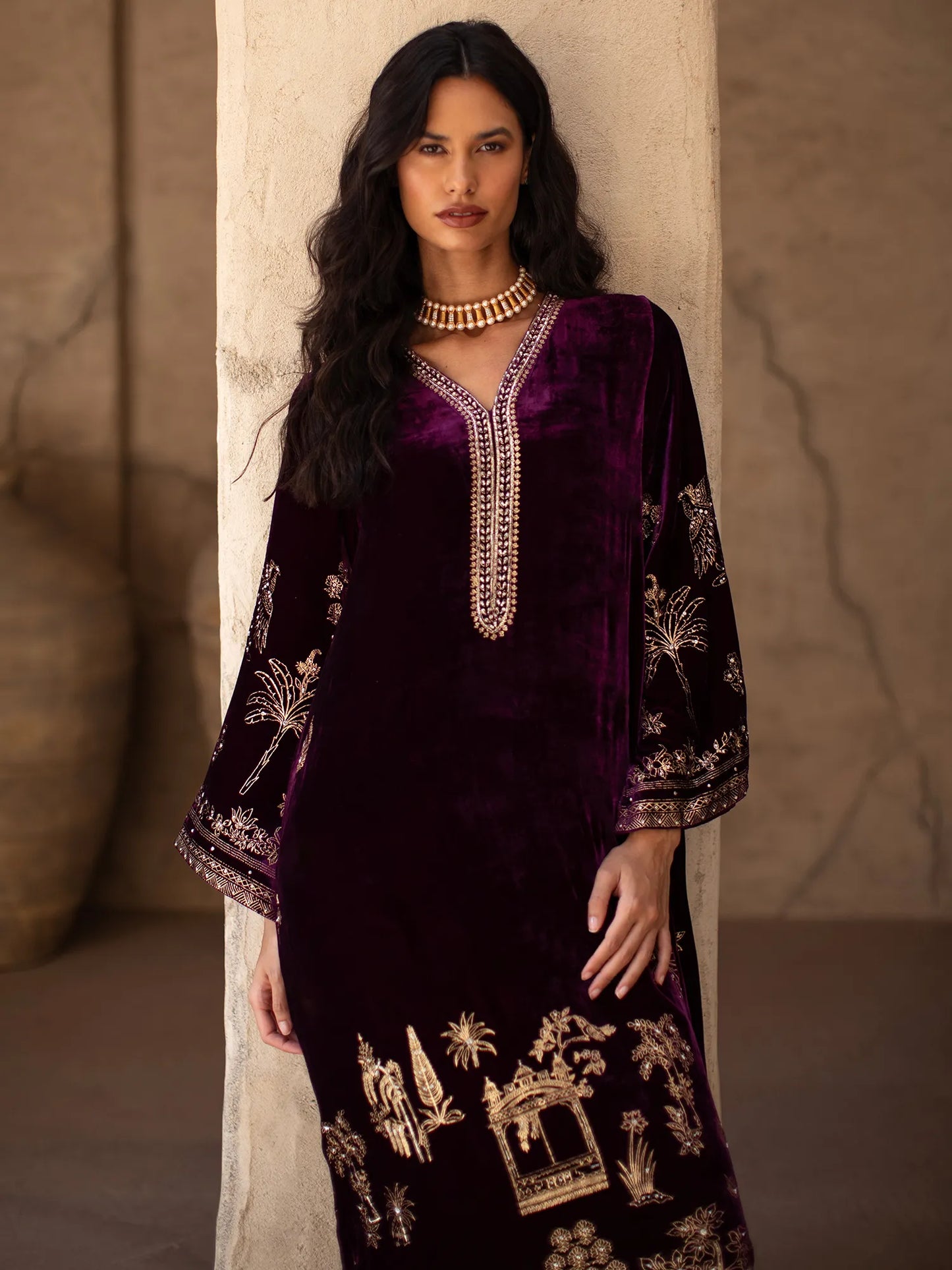 Woman wearing a dark purple dress with intricate gold embroidery against a neutral background