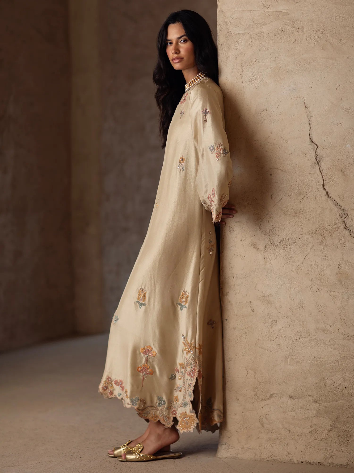 Woman wearing a beige embroidered dress against a textured wall.