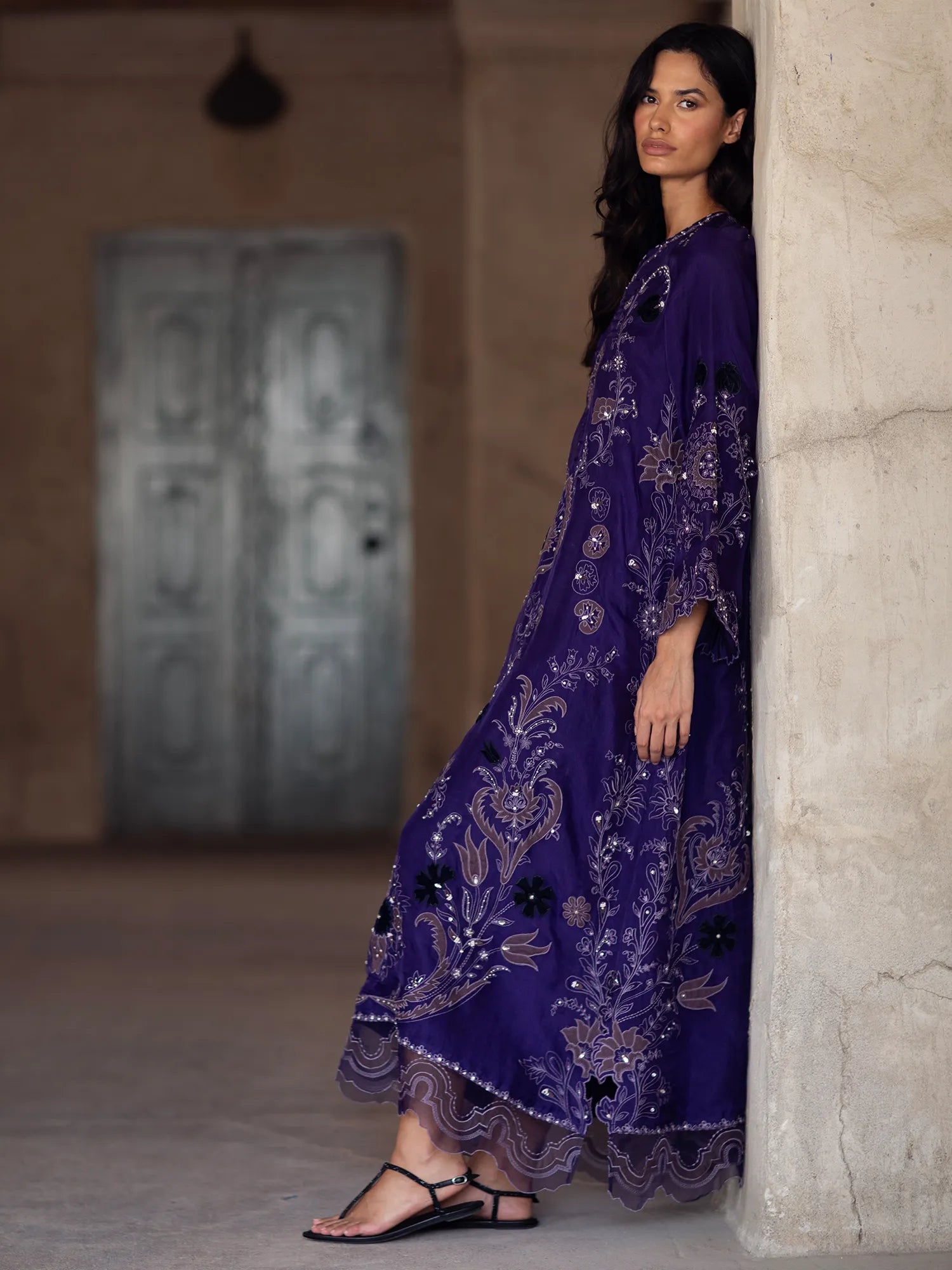 Woman in a long, dark purple embroidered dress standing against a stone wall.