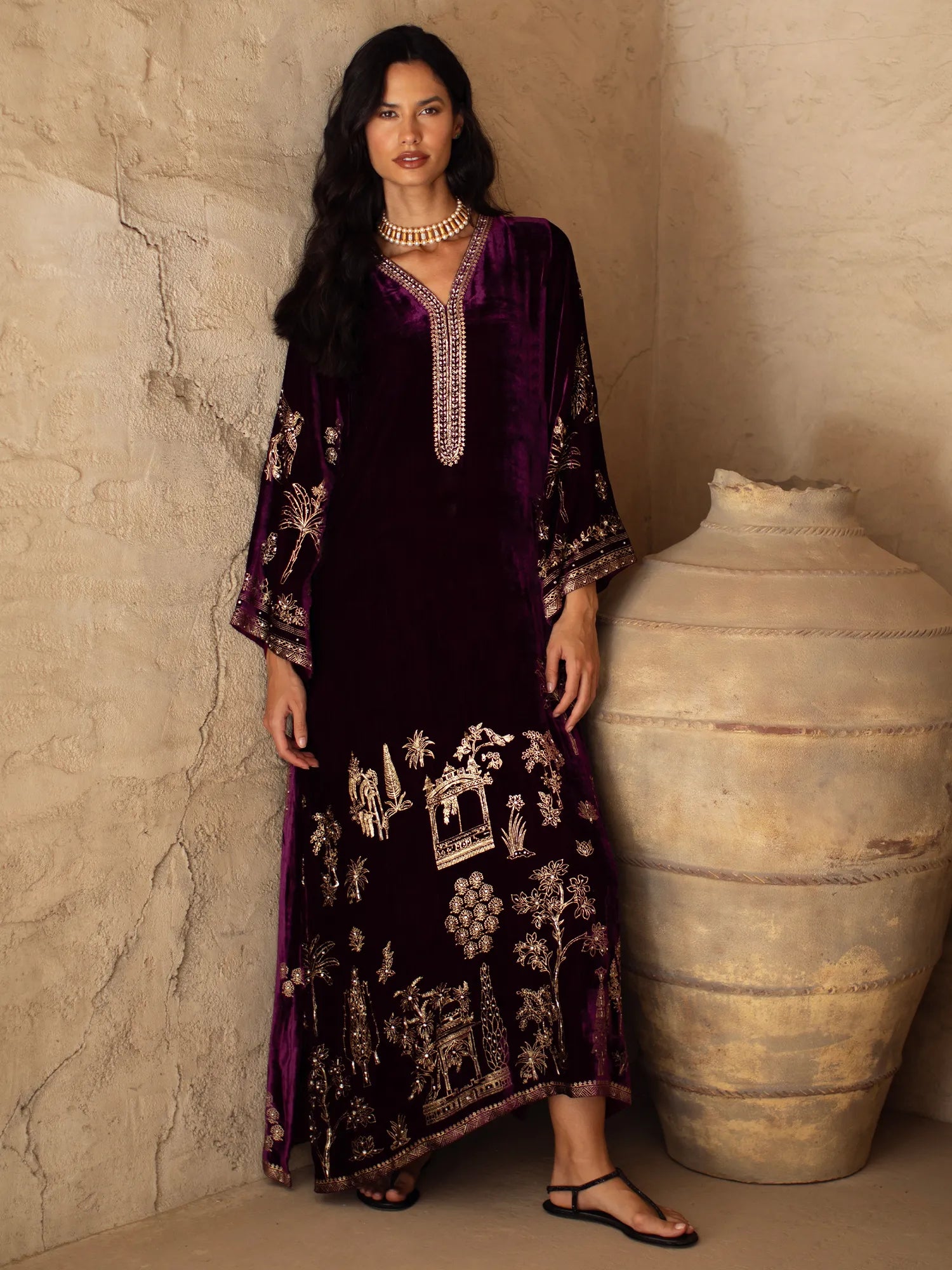 Woman in a dark purple embroidered dress standing next to a large vase against a textured wall.