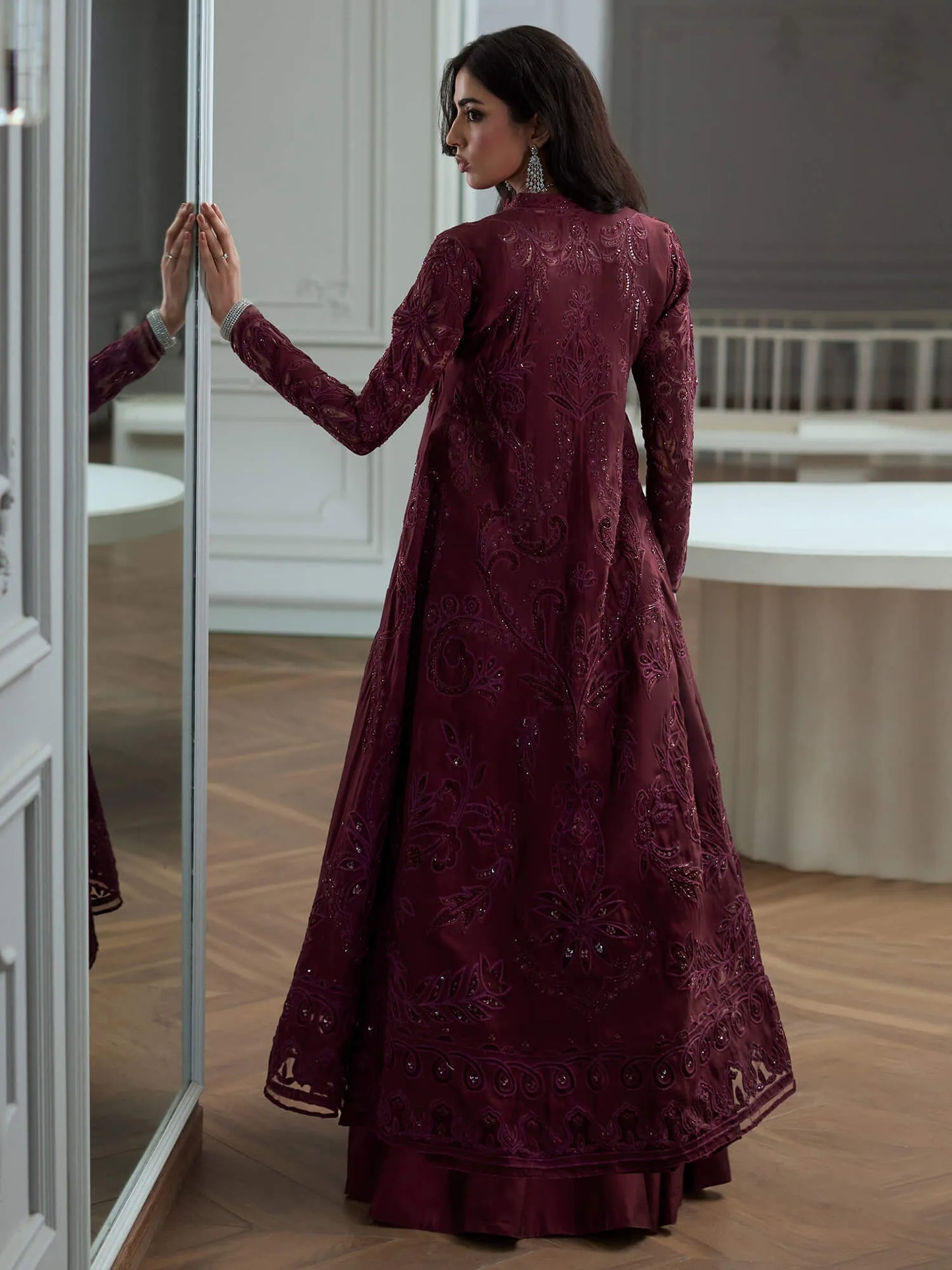 Woman in a burgundy embroidered dress standing in a room with a mirror.