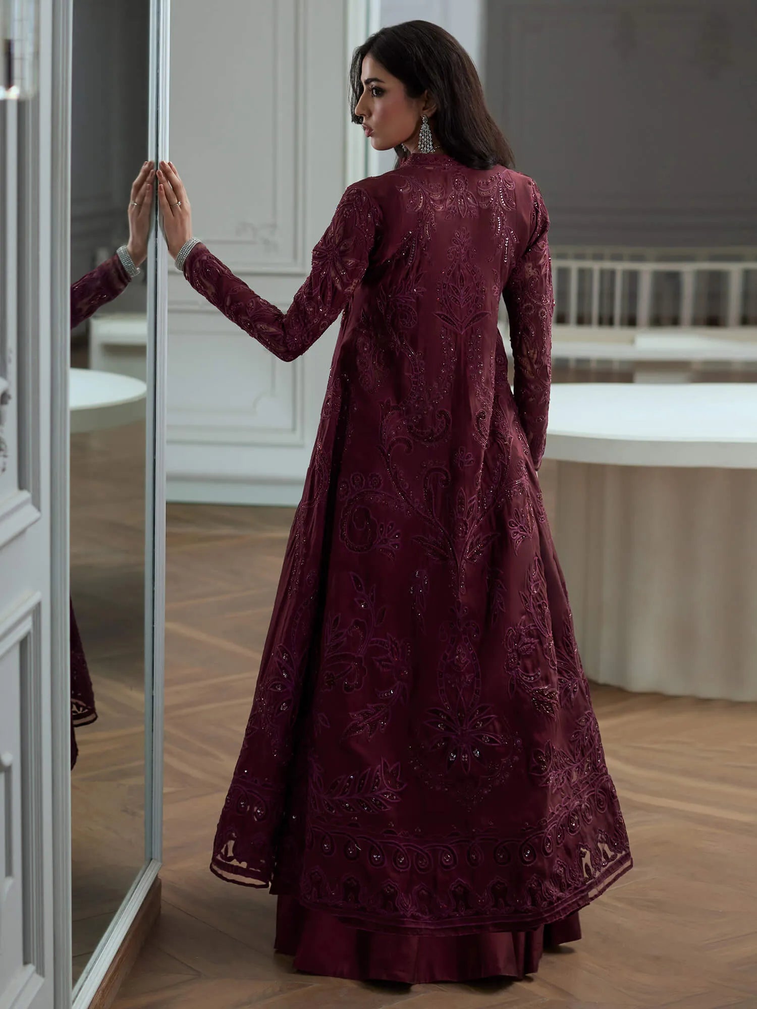 Woman in a burgundy embroidered dress standing in a room with a mirror.
