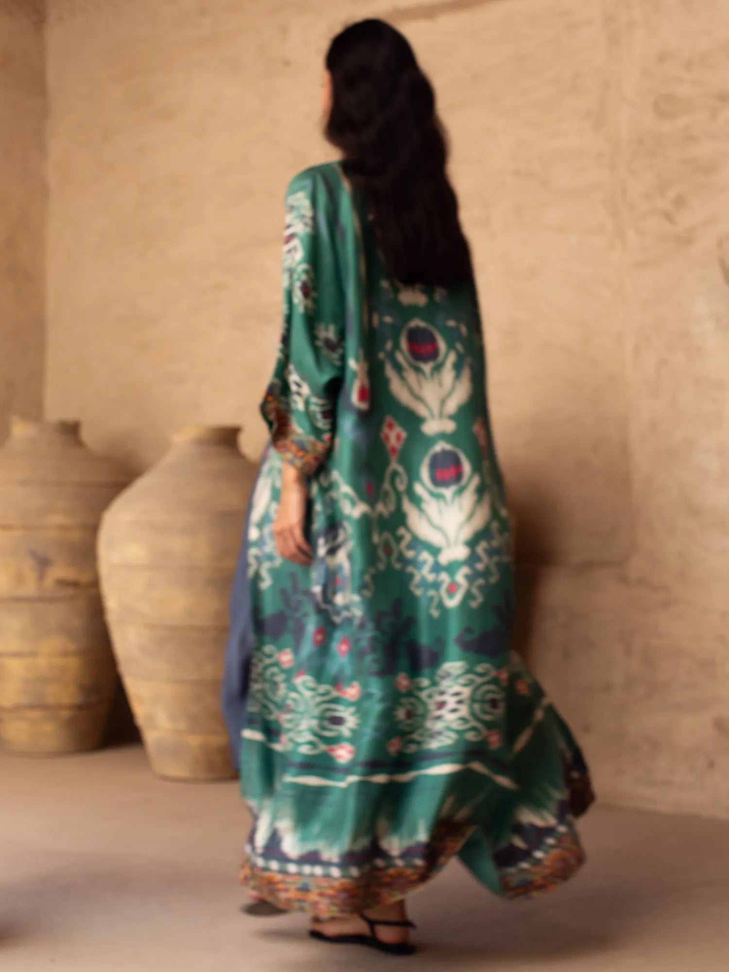 Woman wearing a teal embroidered traditional outfit with a beige wall and large pots in the background