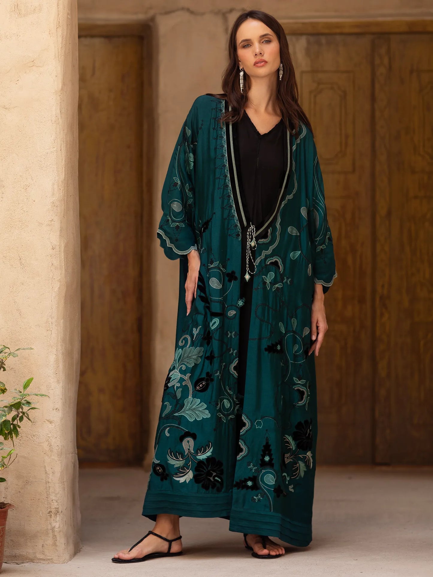 Woman wearing a long teal embroidered dress standing in a warm-toned interior setting.