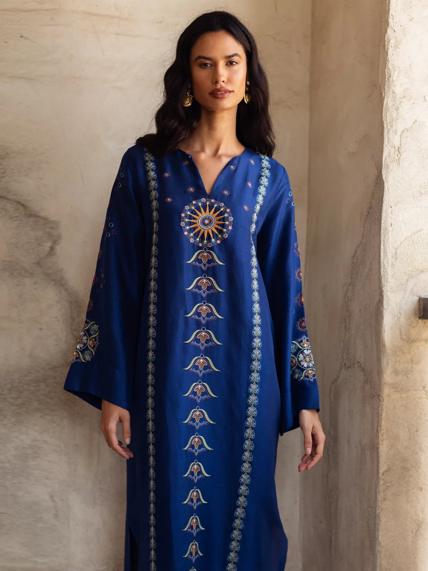 Woman wearing a blue dress with intricate patterns against a neutral wall.