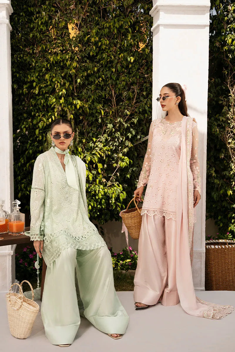 Two women in traditional outfits standing outdoors with greenery and white columns in the background.