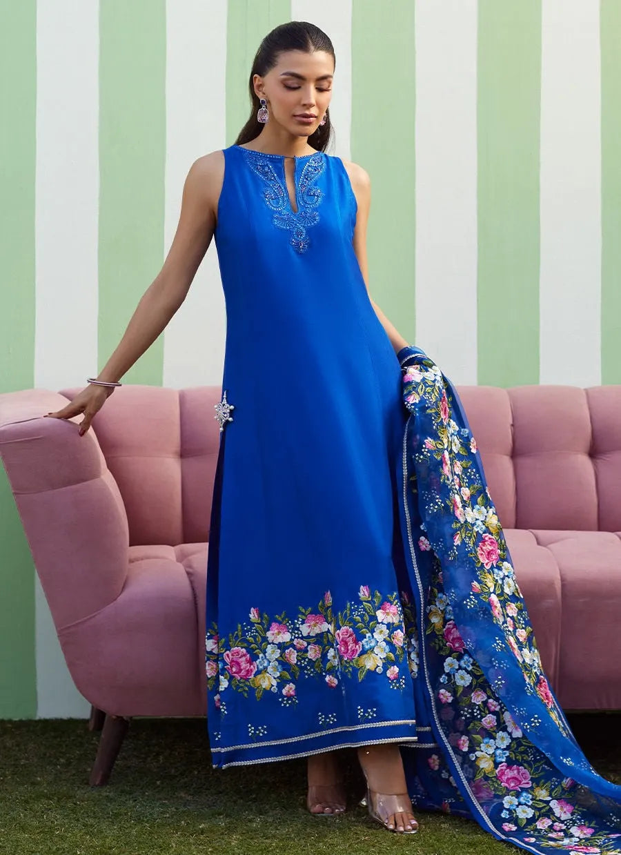Woman in a blue embroidered dress with a floral dupatta standing against a striped wall.