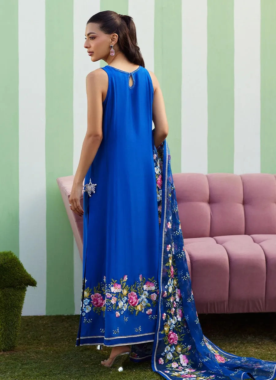 Woman wearing a blue dress with floral embroidery in front of a striped wall and pink sofa.