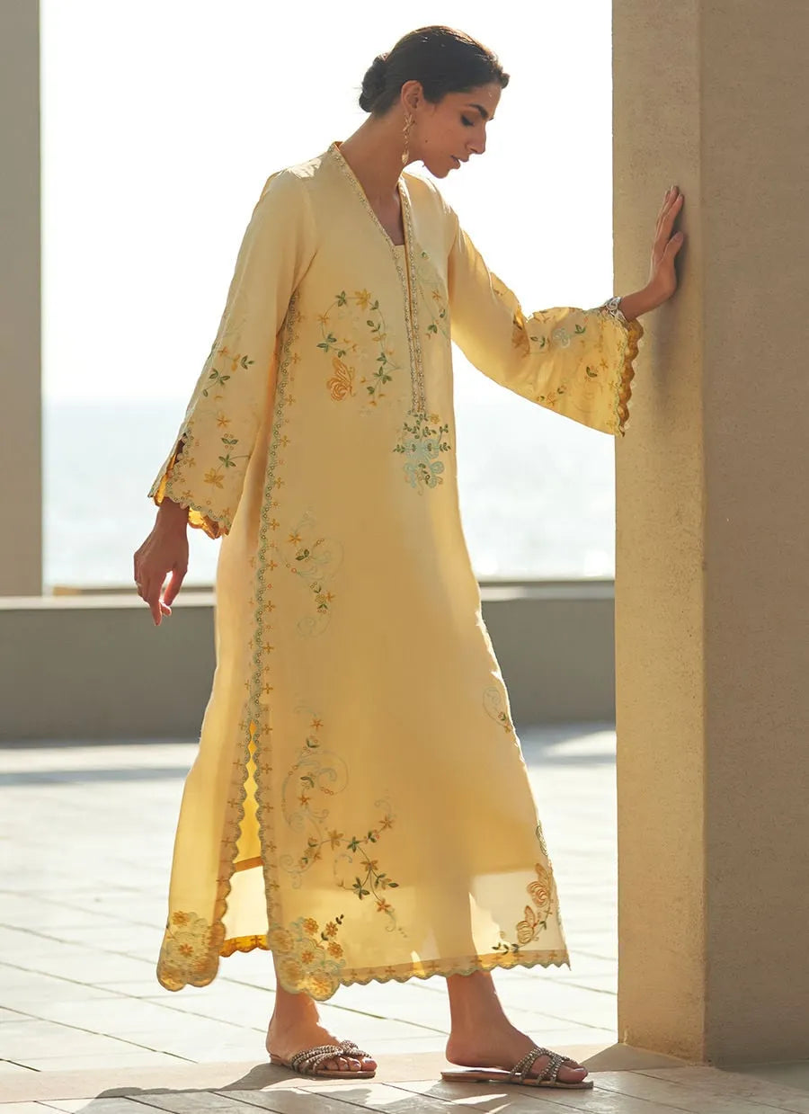 Woman in a yellow embroidered outfit standing by a window with a view of water.