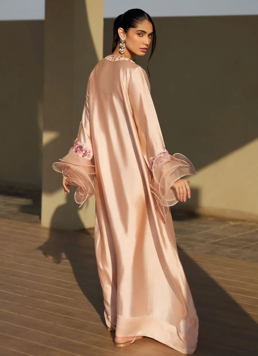 Woman in a long, light pink dress with floral details standing against a neutral background.