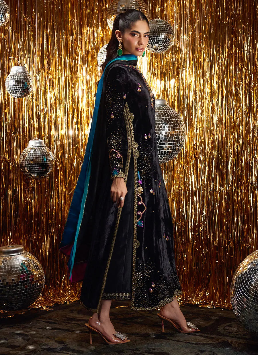 Woman in a black embroidered outfit with a blue dupatta against a glittery gold curtain background.