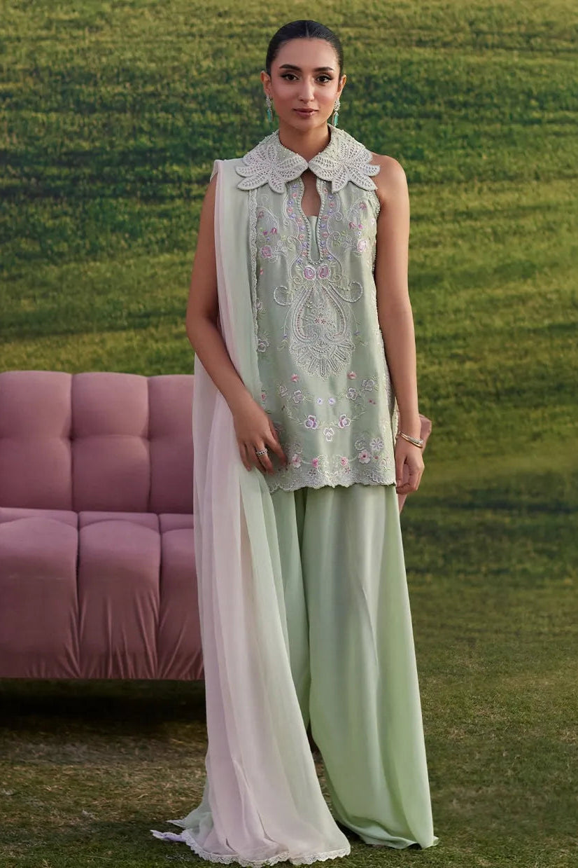 Woman in a light green outfit with floral embroidery standing in a grassy area with a pink couch.