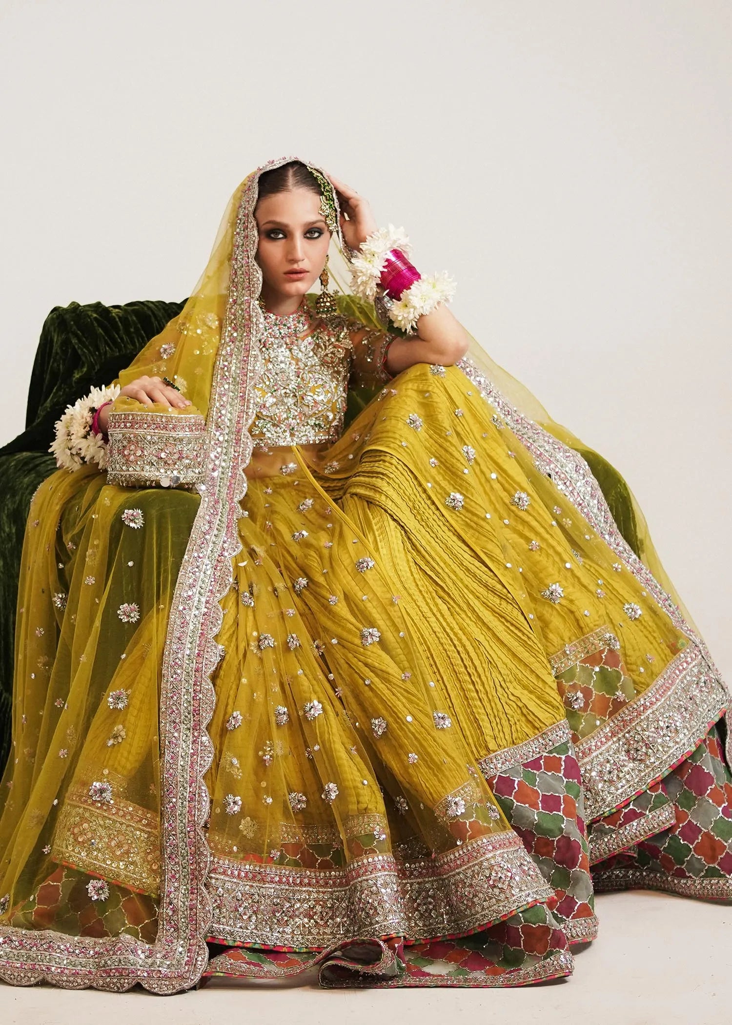 Woman in a traditional yellow and green embroidered outfit with a veil, sitting on a white background.