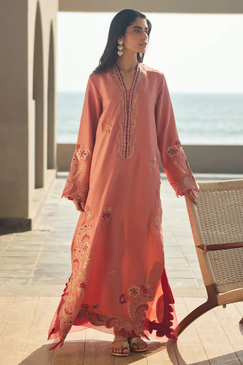 Woman in a peach embroidered dress standing by a wooden chair with a beach view.