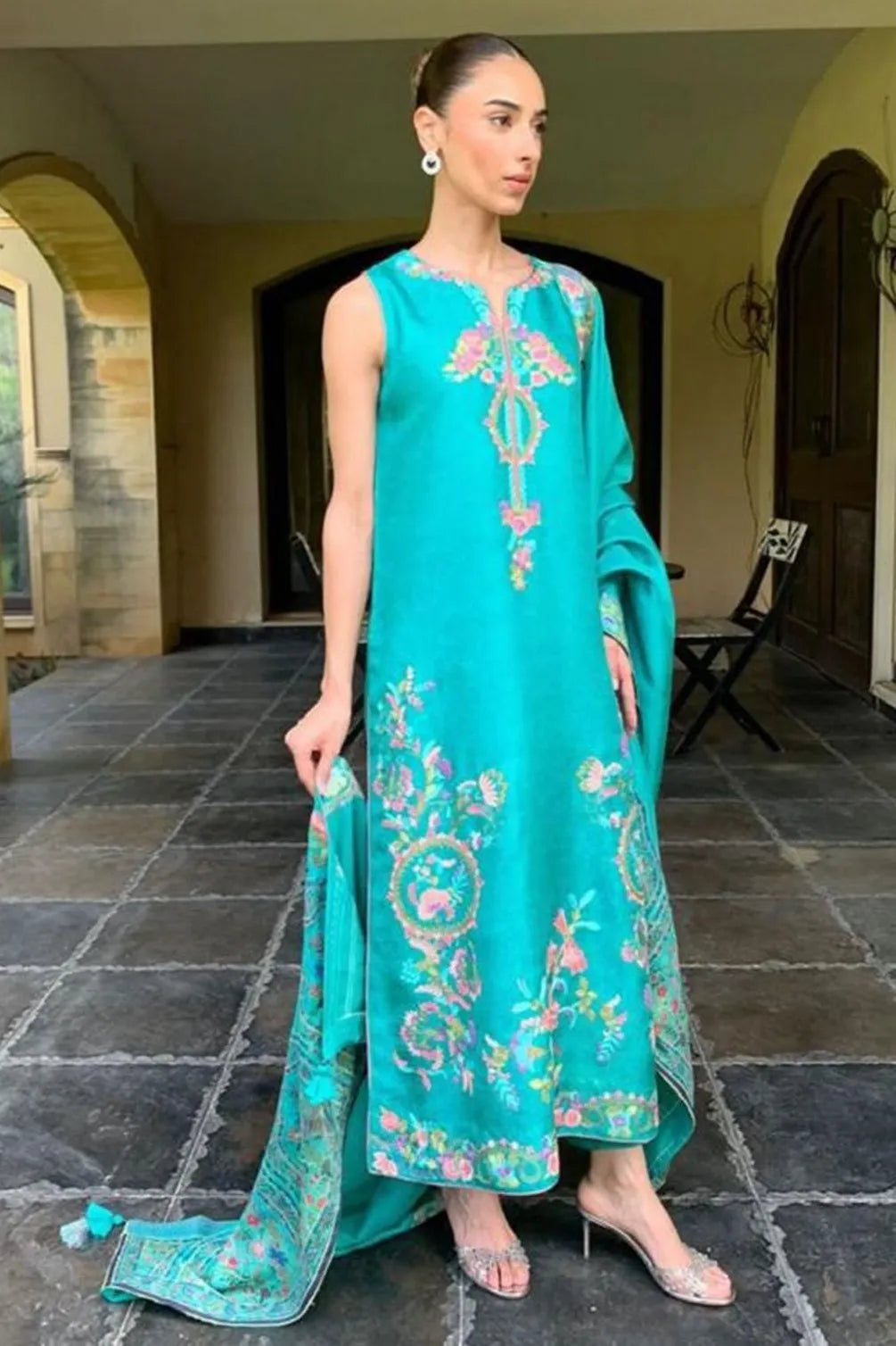 Woman wearing a turquoise embroidered outfit standing on a stone floor.