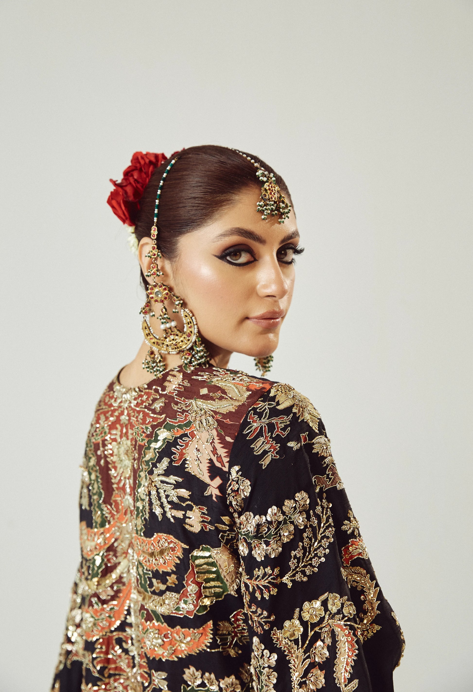 Woman wearing traditional embroidered attire with a red flower in her hair against a plain background