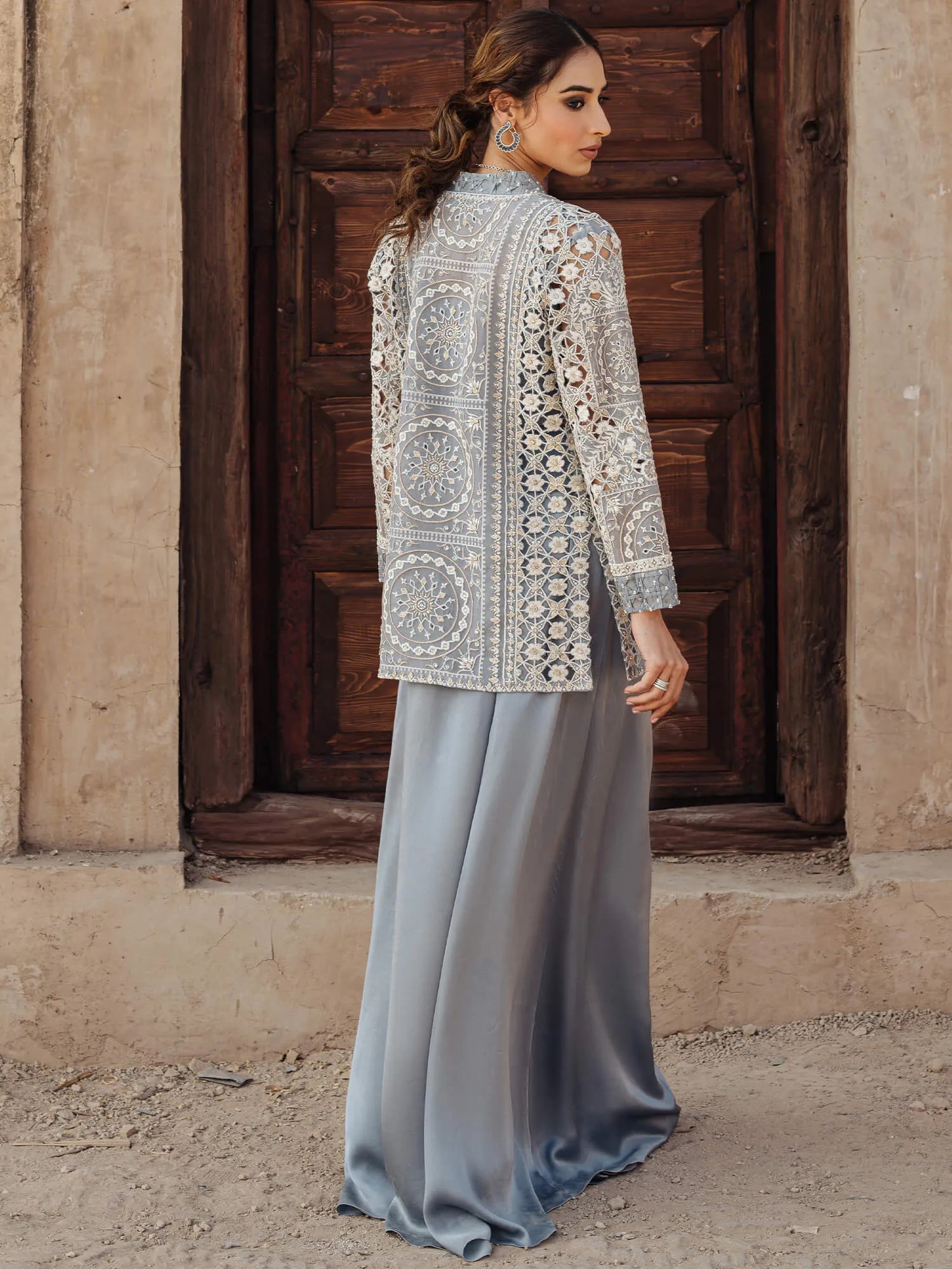 Woman wearing an embroidered top and light blue pants standing in front of a wooden door.